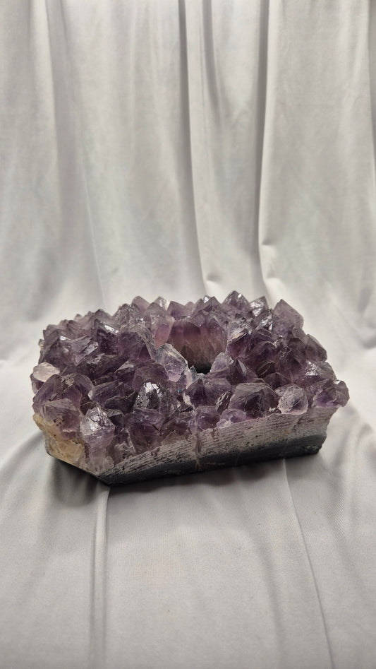 A raw amethyst crystal cluster geode with a natural rough matrix base, displayed against a draped white fabric backdrop.