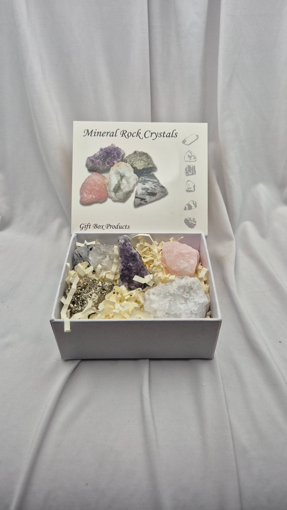 A gift box of mineral and crystal rocks displayed on a draped white fabric background. The open white box contains several raw crystals and rocks nestled in shredded paper filler, with the lid printed with 'Mineral Rock Crystals' and small illustrations.