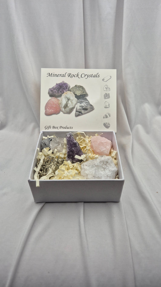 A gift box of mineral and crystal rocks displayed on a draped white fabric background. The open white box contains several raw crystals and rocks nestled in shredded paper filler, with the lid printed with 'Mineral Rock Crystals' and small illustrations.