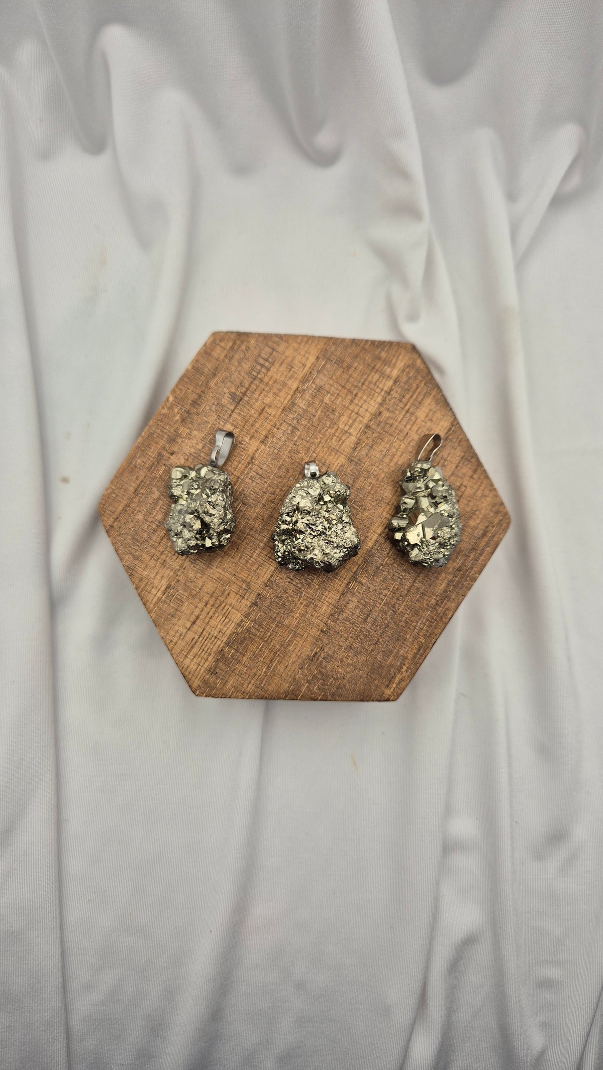 Three small pyrite pendants with silver-toned loops are displayed on a hexagonal wooden block over a draped white fabric background.