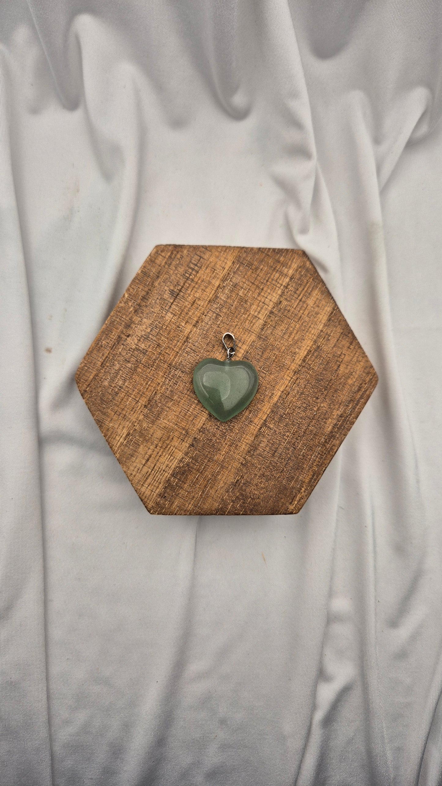 A small heart-shaped green stone pendant with a silver bail, displayed on a hexagonal wooden block over a draped white fabric background.