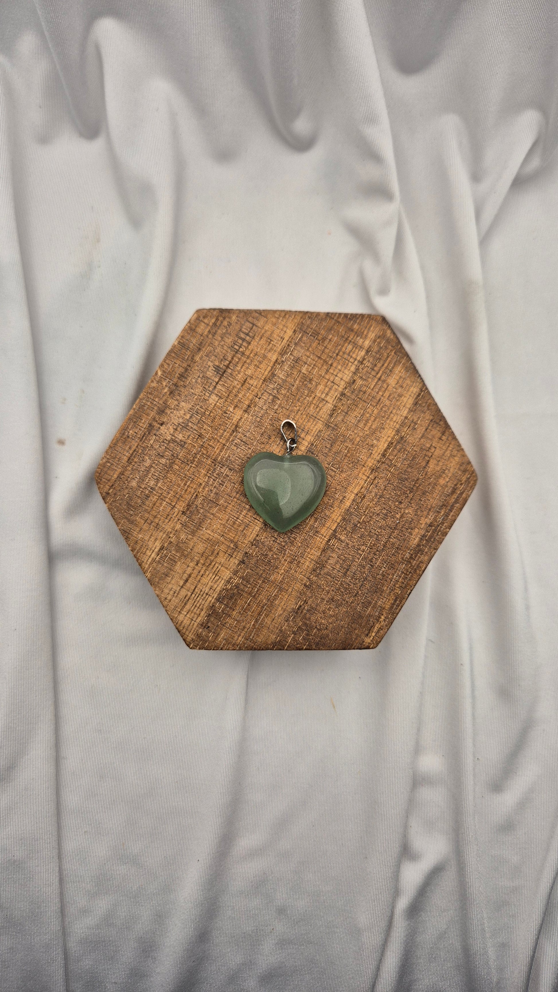 A small heart-shaped green stone pendant with a silver bail, displayed on a hexagonal wooden block over a draped white fabric background.