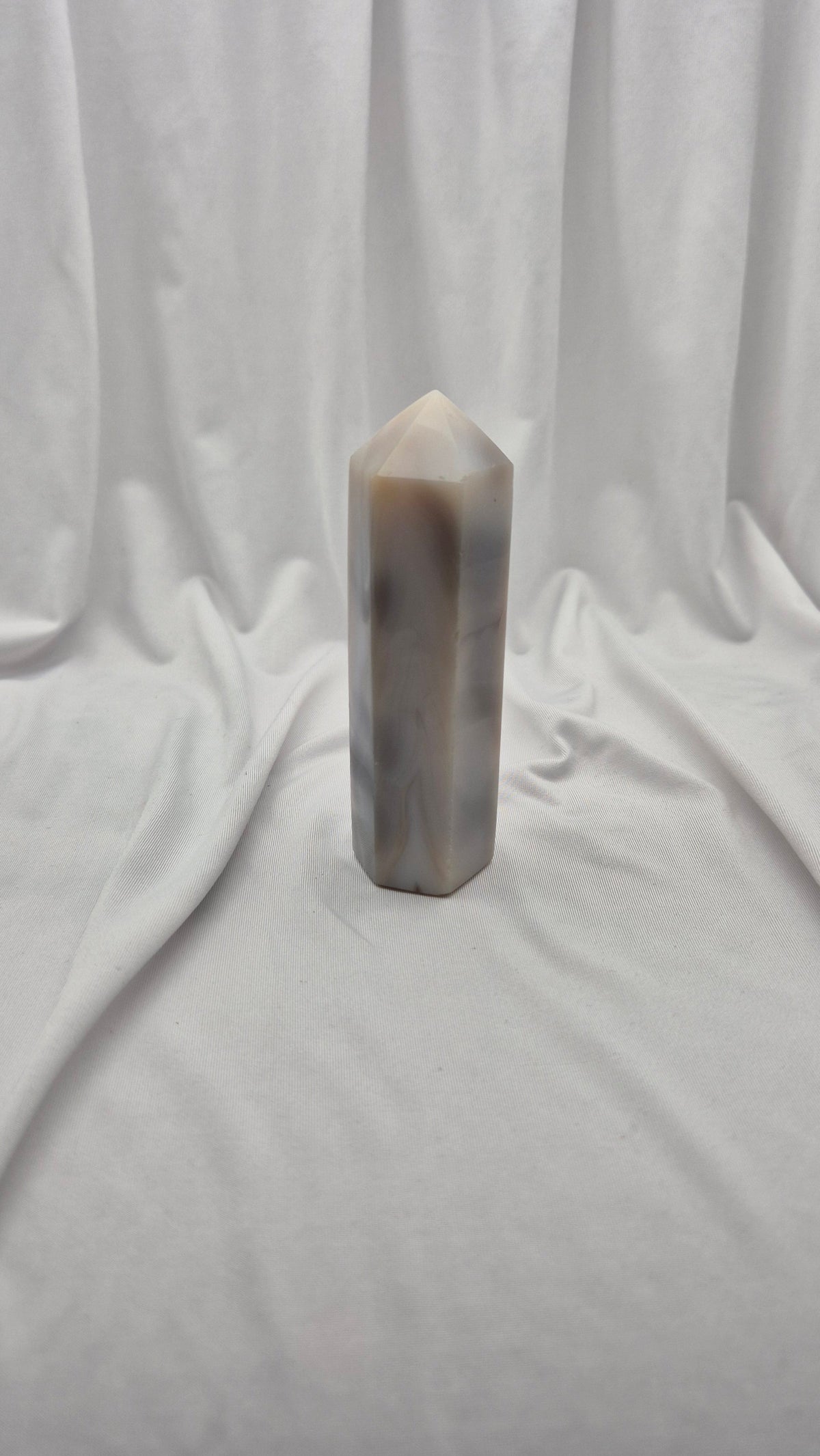 A polished natural quartz crystal tower with a pointed top, featuring natural beige and gray banded patterns, standing upright on draped white fabric.