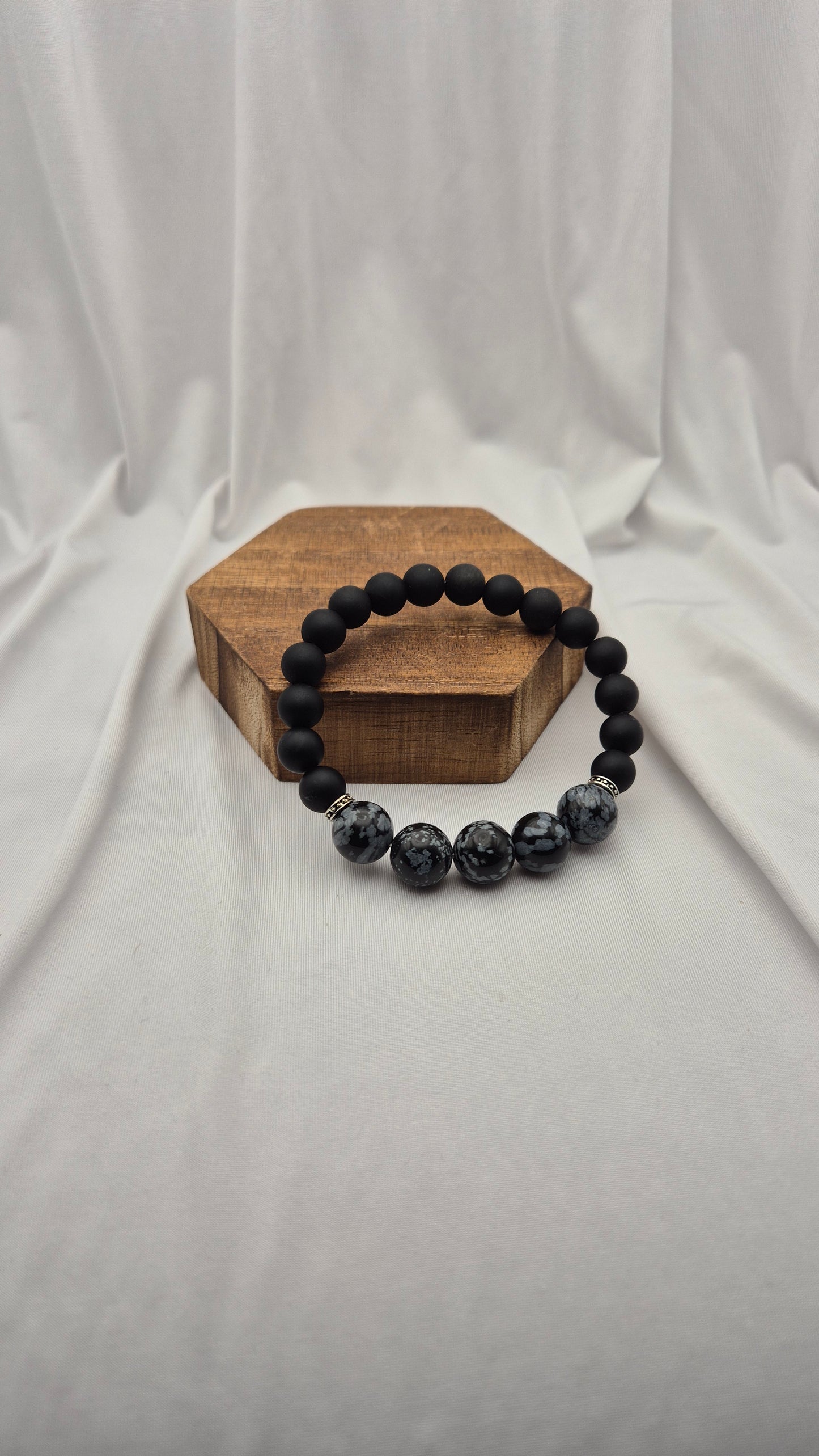 A beaded stretch bracelet with matte black beads and three marbled dark gray and black beads, displayed on a small hexagonal wooden block against a draped white fabric background.