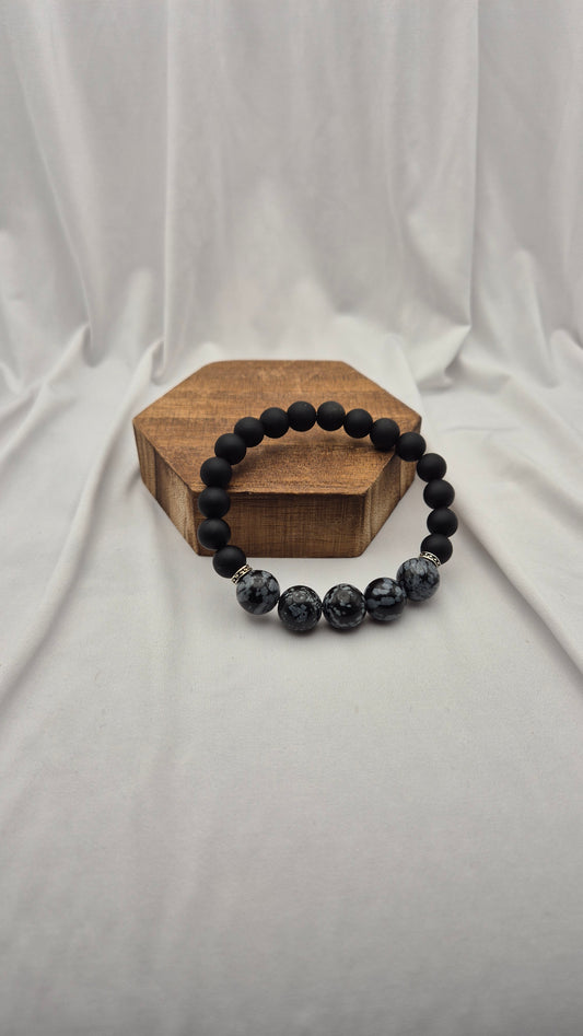 A beaded stretch bracelet with matte black beads and three marbled dark gray and black beads, displayed on a small hexagonal wooden block against a draped white fabric background.