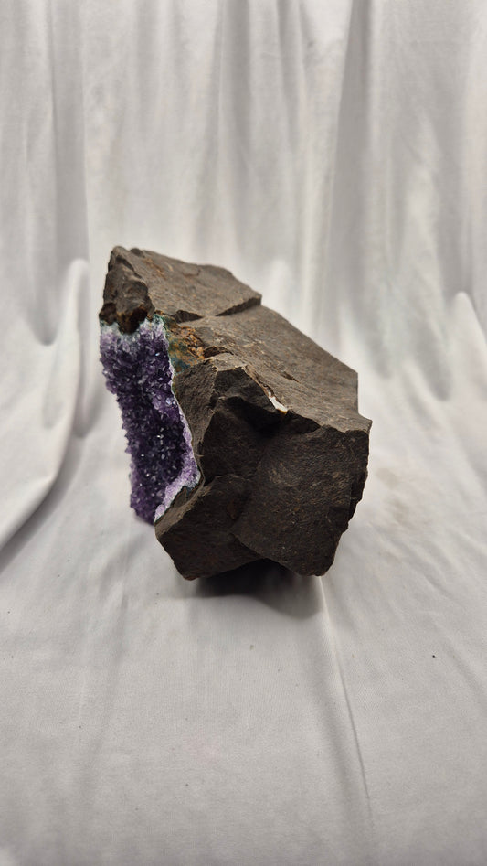 A raw amethyst geode specimen with a dark brown to black volcanic matrix, photographed on a draped white fabric background.