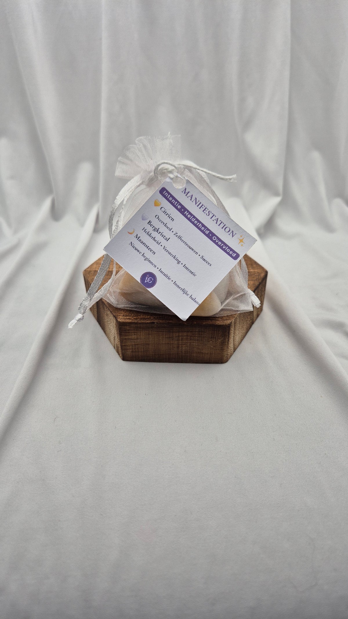A small white sheer drawstring pouch with a printed tag sits on a hexagonal wooden tray against a draped white fabric backdrop.