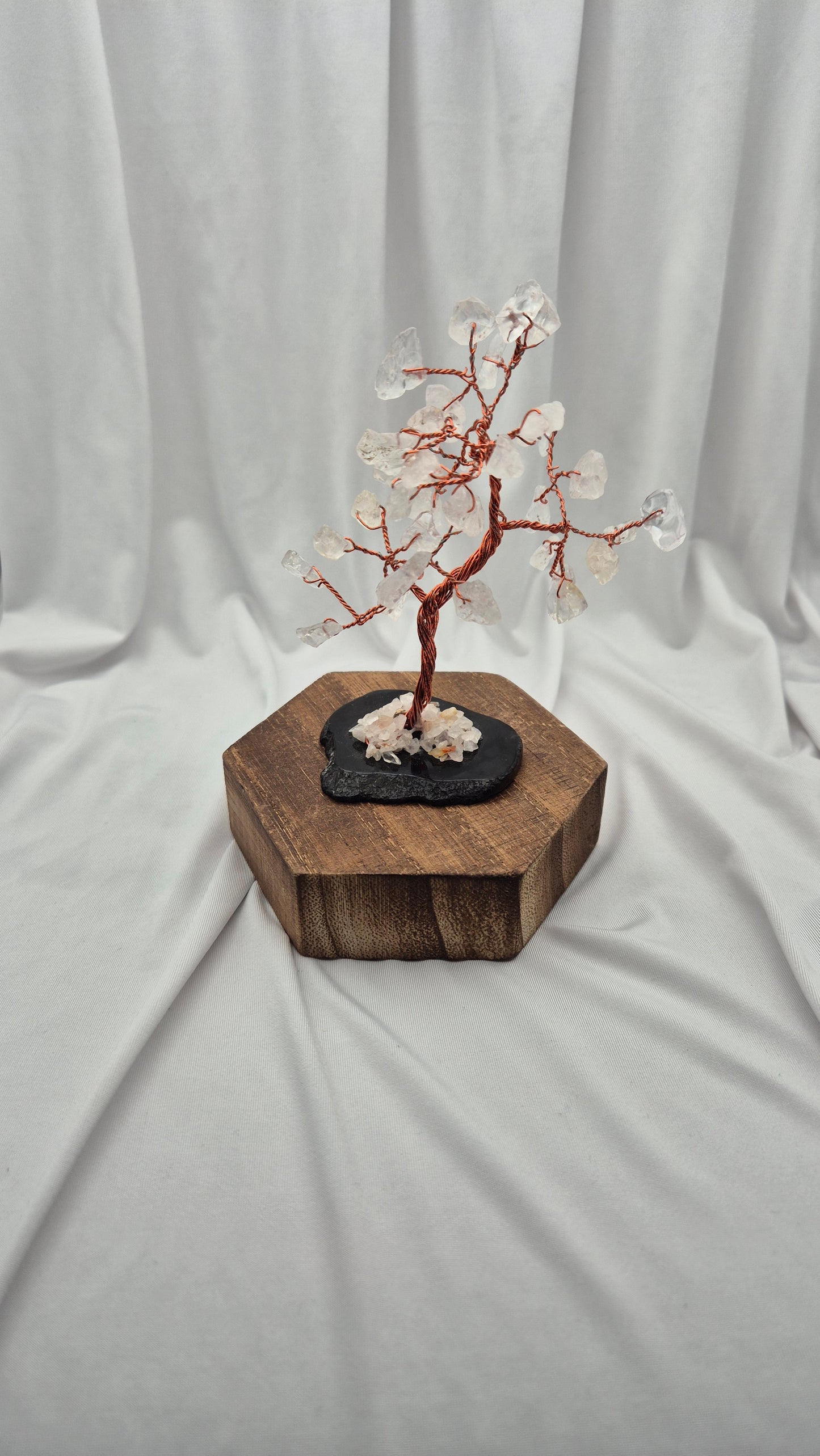 A crystal tree sculpture with a reddish-brown wire trunk and white crystal blossoms, displayed on a black stone base atop a hexagonal wooden block against a draped white fabric backdrop.