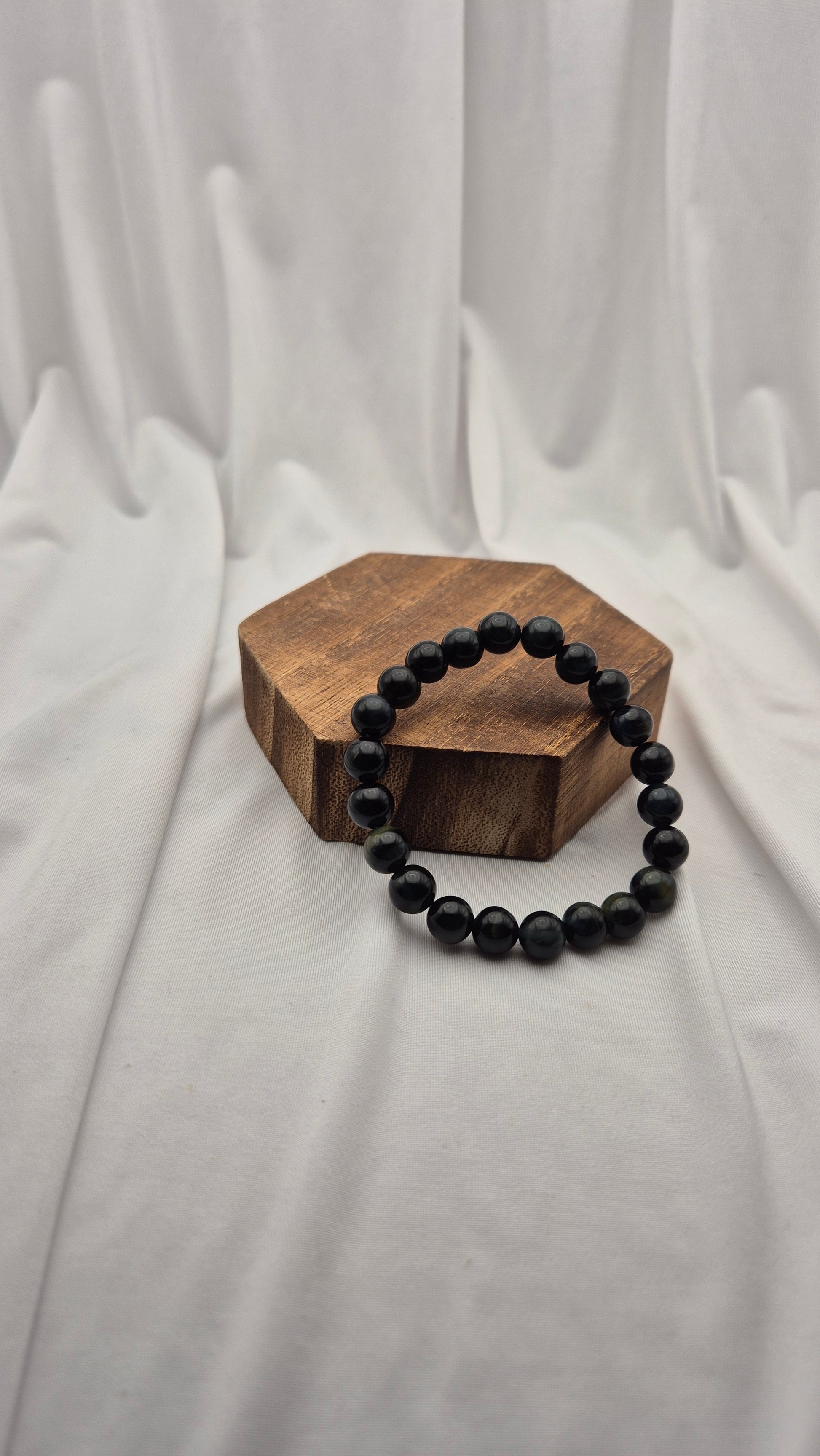 A beaded bracelet made of matte black round beads, displayed on a small hexagonal wooden block against a draped white fabric backdrop.