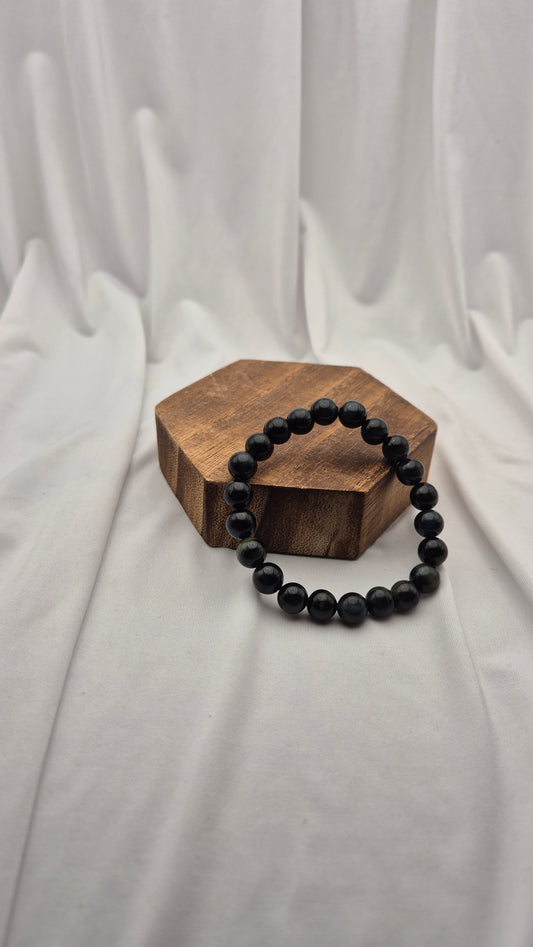 A beaded bracelet made of matte black round beads, displayed on a small hexagonal wooden block against a draped white fabric backdrop.