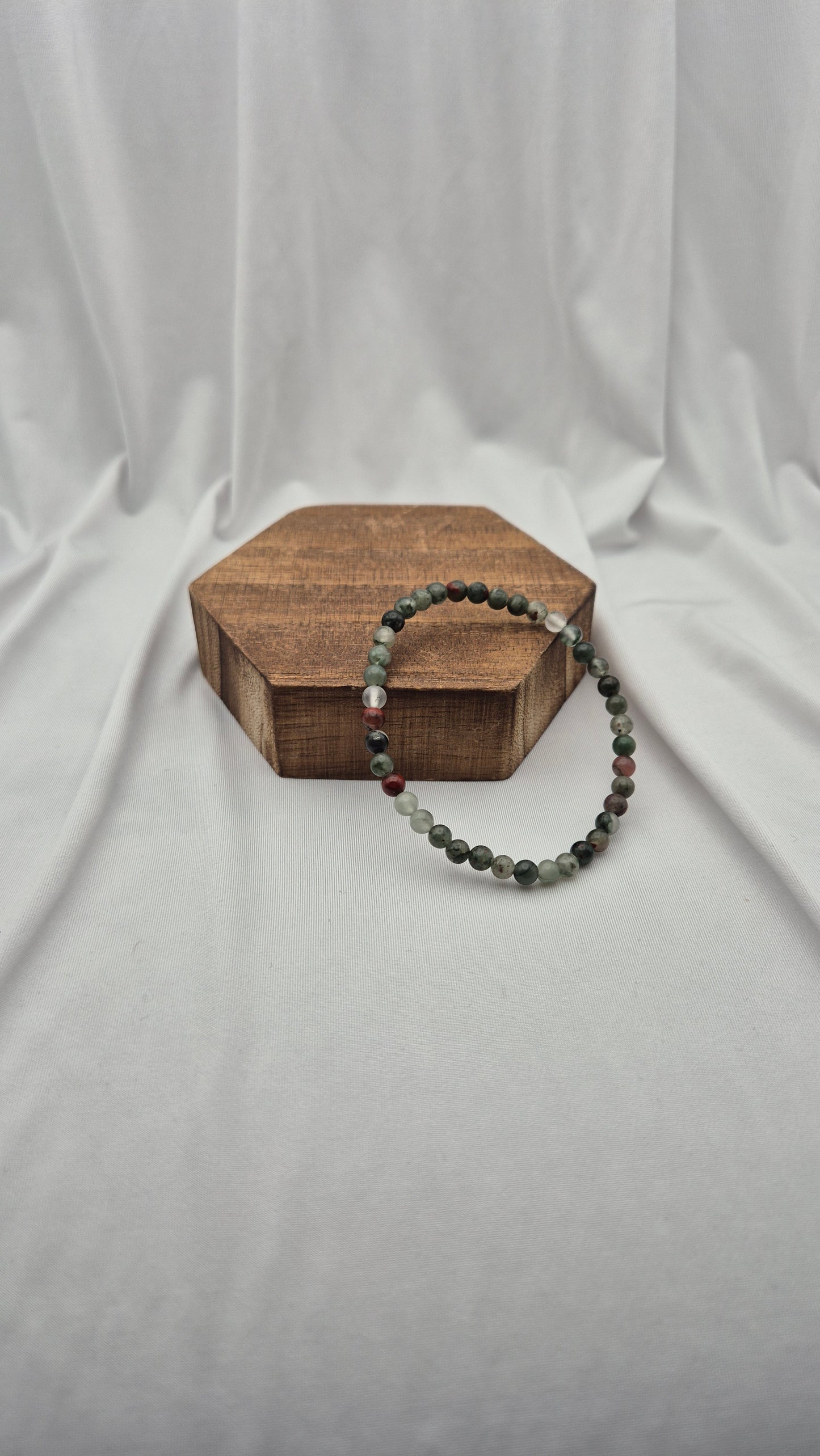 A beaded stretch bracelet with dark green, gray, and red beads draped over a hexagonal wooden block, set against a draped white fabric background.