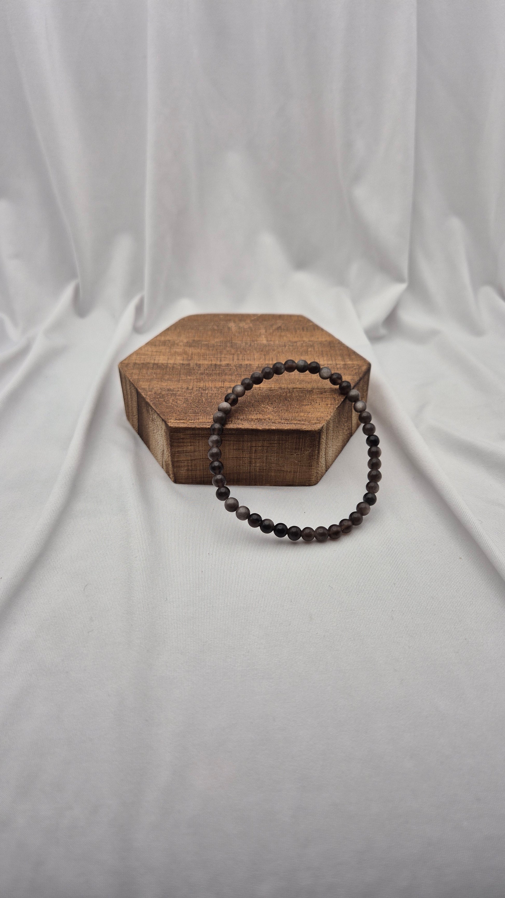 A dark brown beaded bracelet with small, polished round beads, displayed on a hexagonal wooden block against a draped white fabric backdrop.