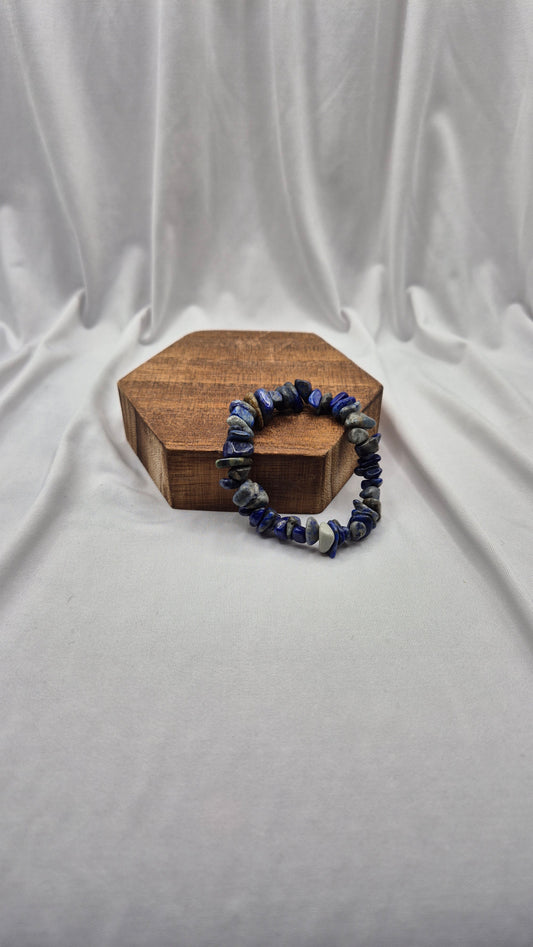 A beaded stretch bracelet made of dark blue and gray stone chips, displayed on a small hexagonal wooden box against a draped white fabric backdrop.