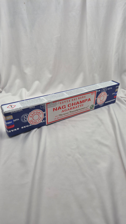 A long rectangular box of Nag Champa Agarbatti (incense sticks) placed diagonally on a wrinkled white fabric background. The packaging is primarily dark blue with a central red label and white text.