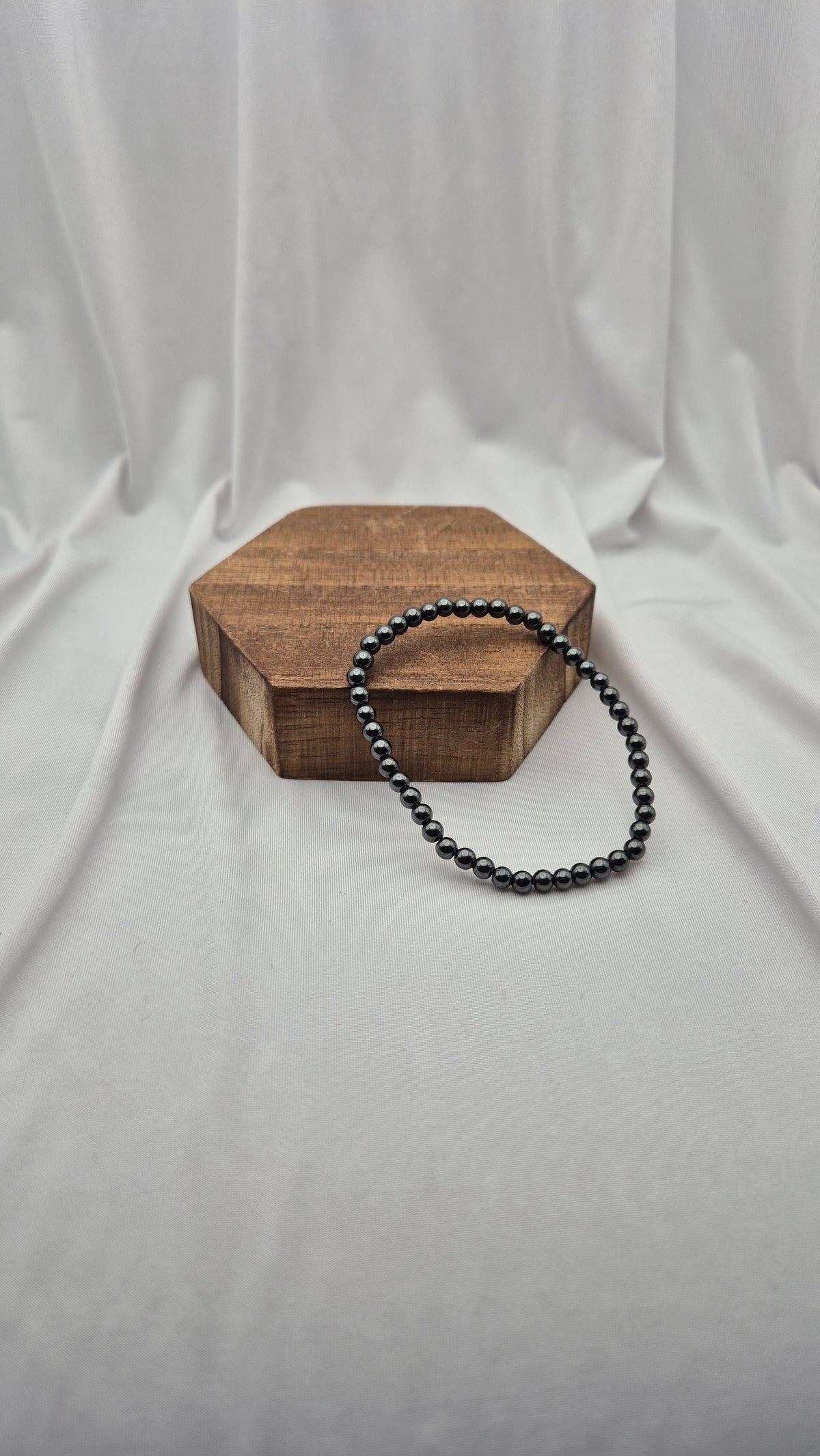 A black beaded bracelet draped over a hexagonal wooden block, set against a draped white fabric backdrop.