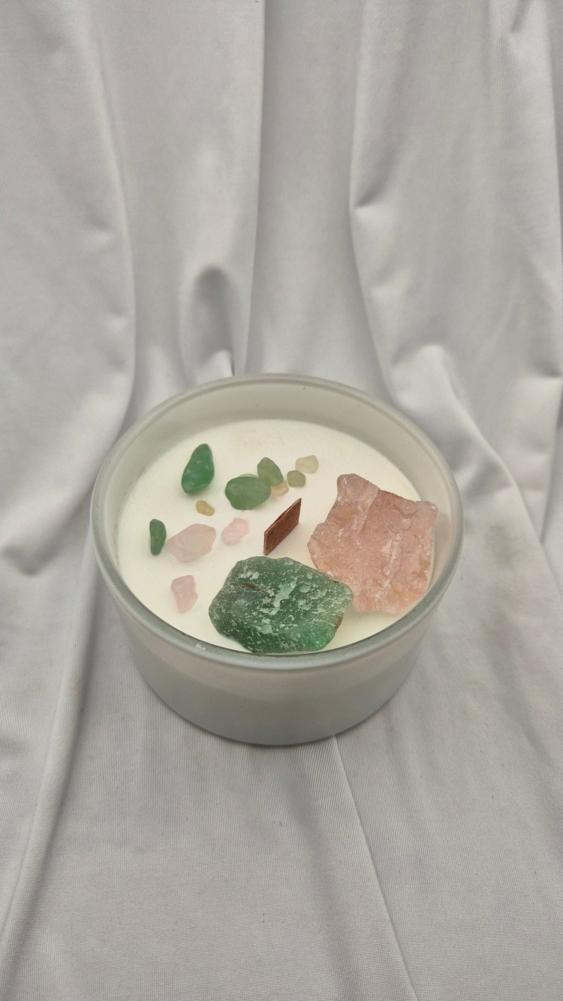 A glass jar candle filled with white wax and topped with various raw crystals, including green, pink, and brown stones, set against a draped white fabric background.