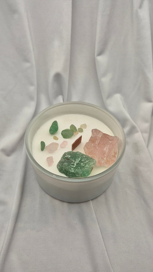 A glass jar candle filled with white wax and topped with various raw crystals, including green, pink, and brown stones, set against a draped white fabric background.