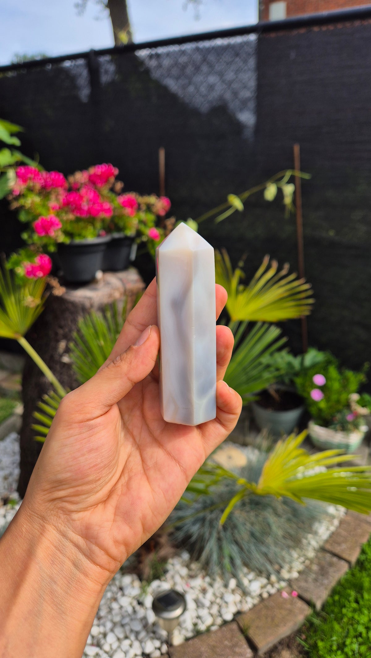 A hand holding a polished white and gray marble point stone against a blurred outdoor garden background with potted plants and a black fence.