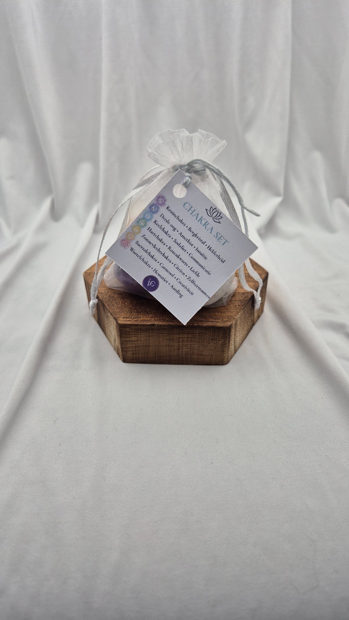 A small clear mesh gift bag tied with a sheer ribbon, resting on a hexagonal wooden block. A printed tag attached to the bag reads 'CHANEL SET' with additional text below, photographed against draped white fabric.