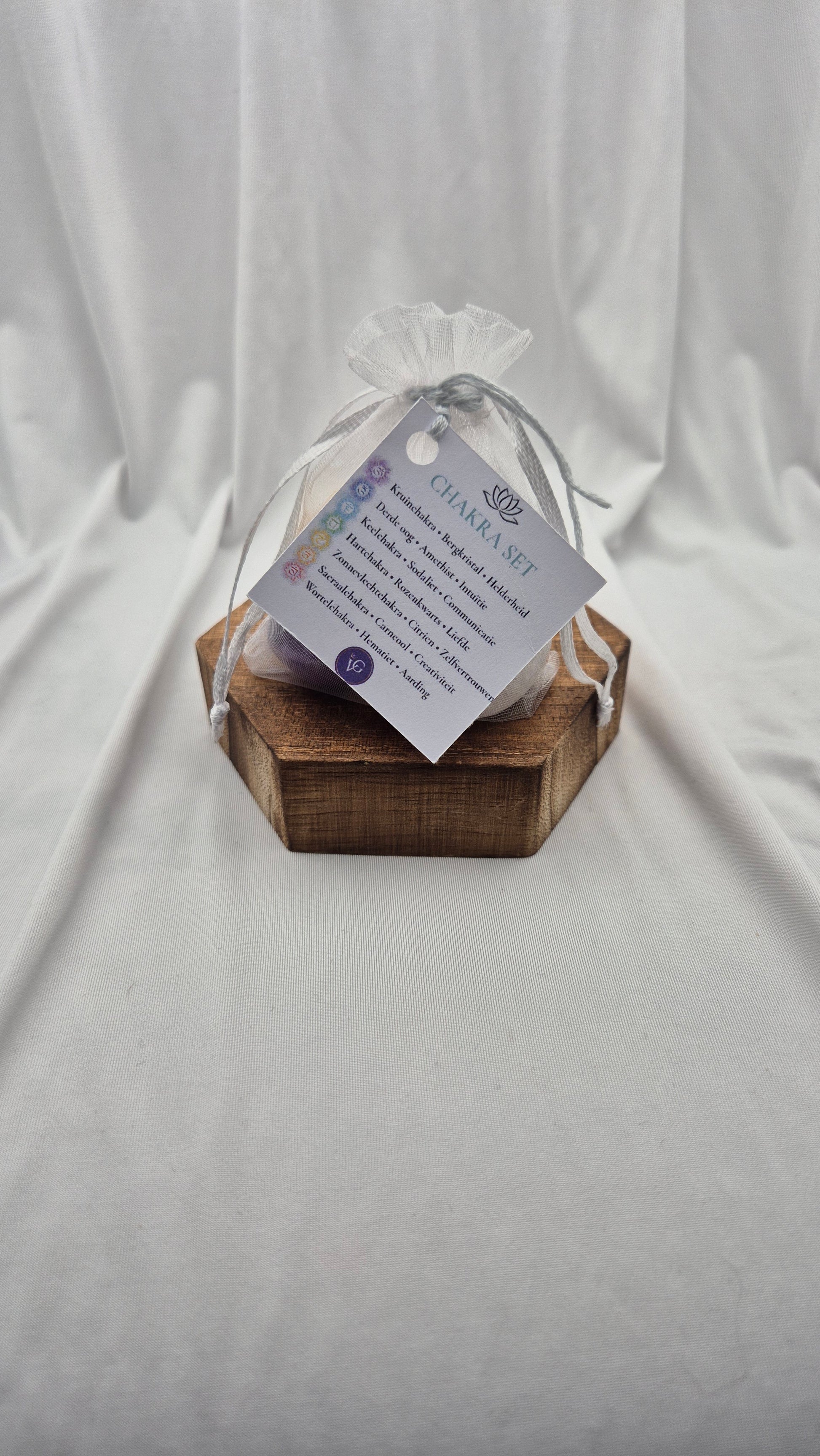 A small clear mesh gift bag tied with a sheer ribbon, resting on a hexagonal wooden block. A printed tag attached to the bag reads 'CHANEL SET' with additional text below, photographed against draped white fabric.