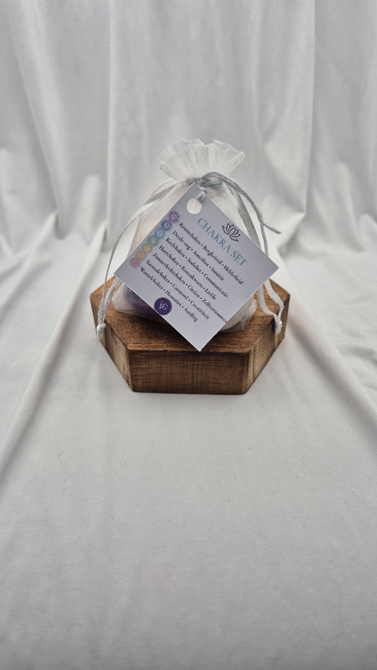 A small clear mesh gift bag tied with a sheer ribbon, resting on a hexagonal wooden block. A printed tag attached to the bag reads 'CHANEL SET' with additional text below, photographed against draped white fabric.