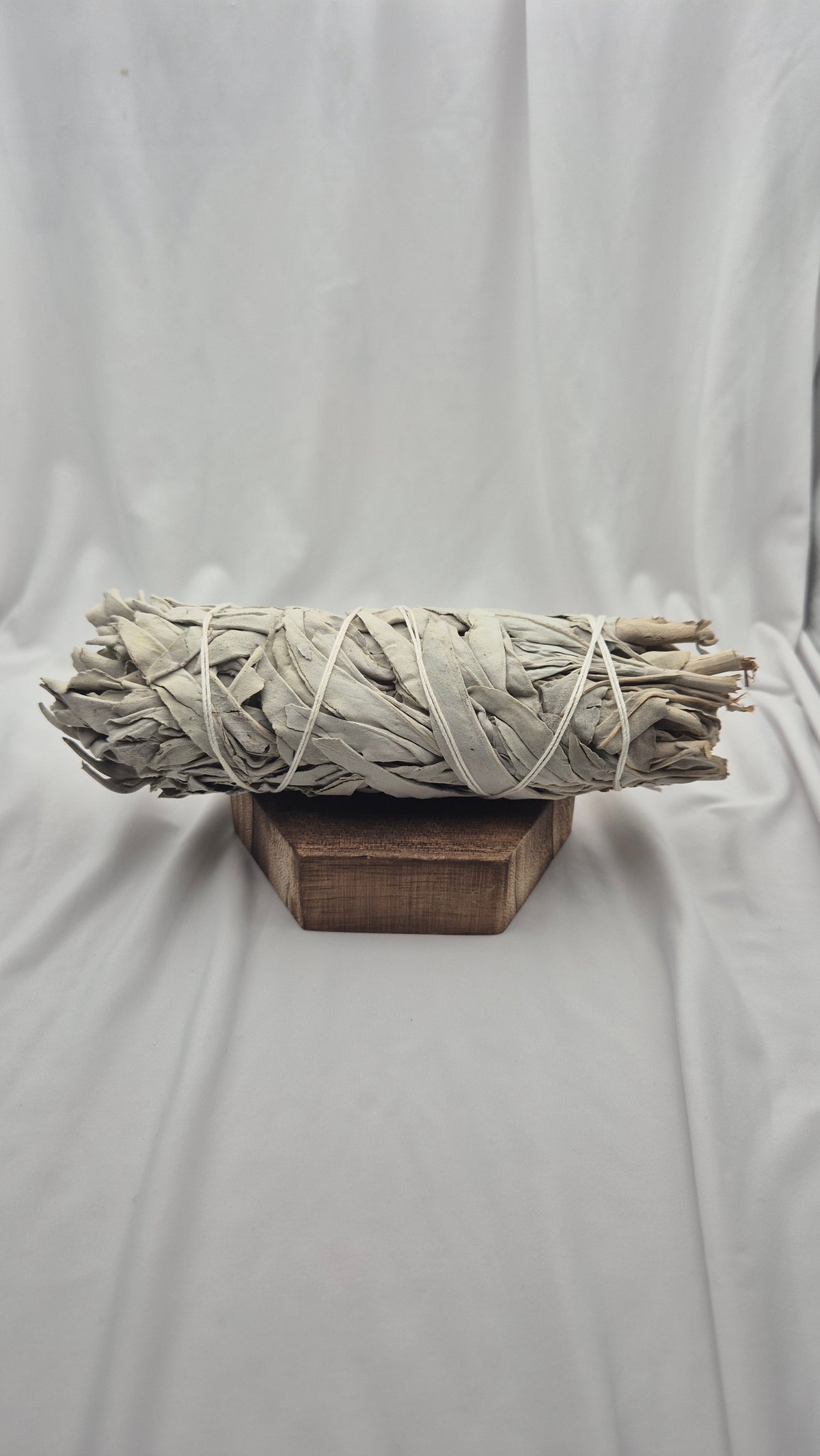 A bundle of dried white sage smudge sticks wrapped in natural cord, displayed on a small wooden block against a draped white fabric backdrop.
