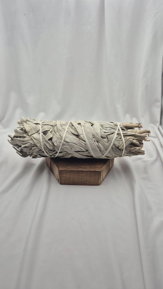 A bundle of dried white sage smudge sticks wrapped in natural cord, displayed on a small wooden block against a draped white fabric backdrop.