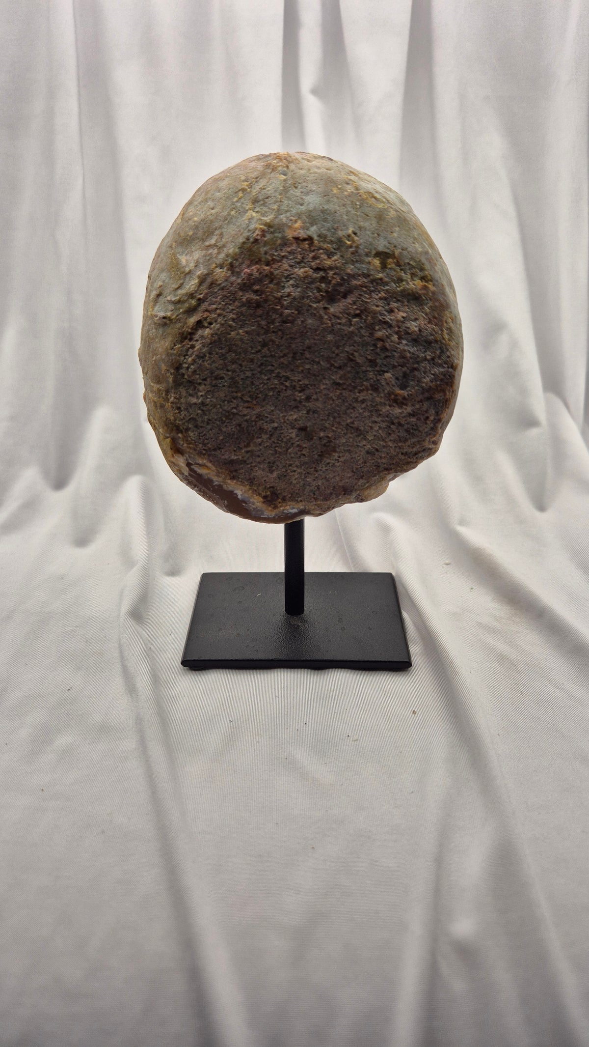 A large, oval-shaped natural stone or rock specimen displayed on a black metal stand against a draped white fabric backdrop.