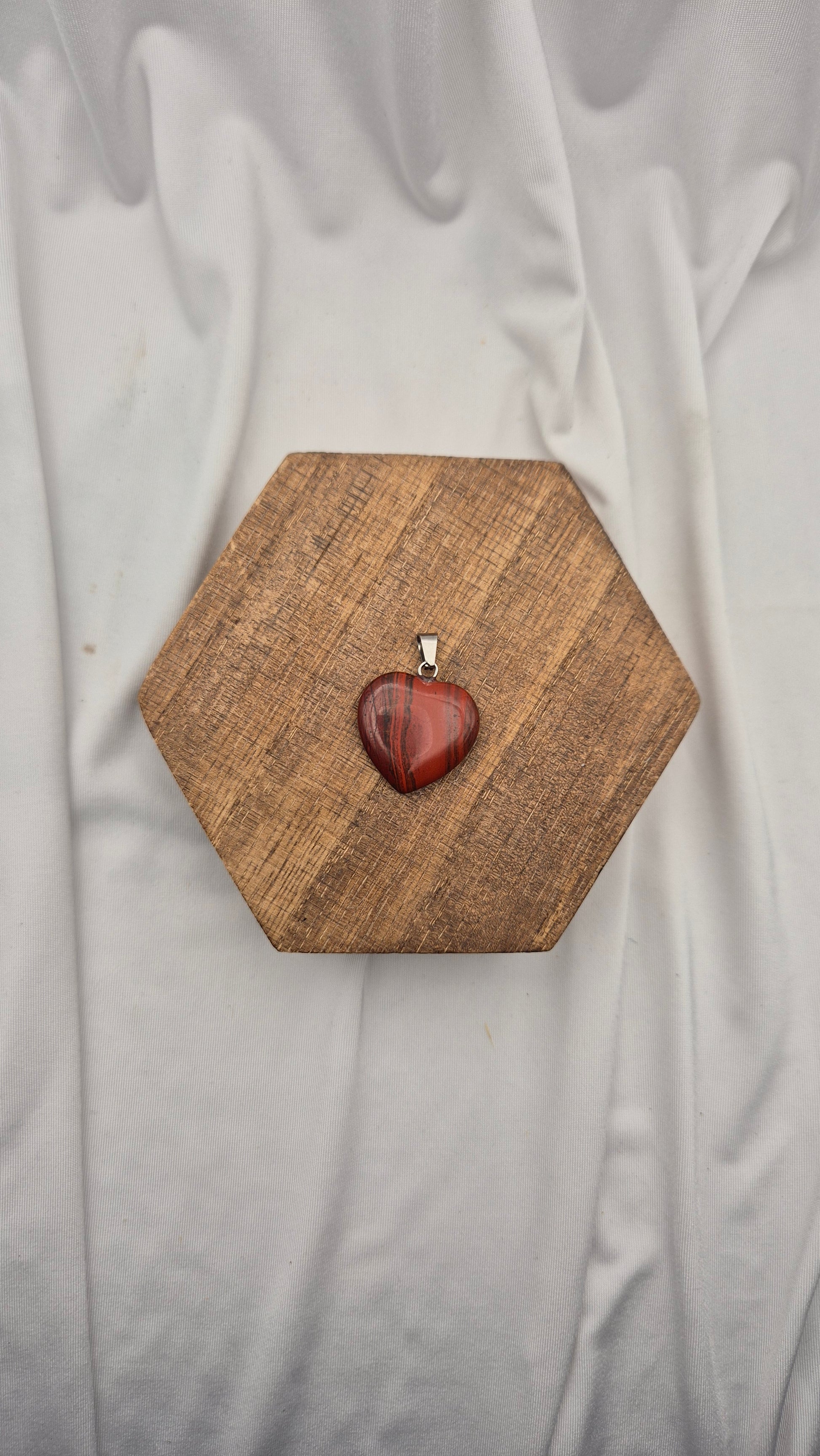 A heart-shaped red stone pendant with a silver bail, displayed on a hexagonal wooden block over a draped white fabric background.