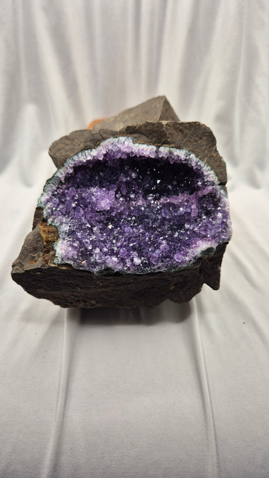 A raw amethyst geode specimen with a dark brown to black volcanic matrix, showing a vibrant purple and lavender crystalline interior, photographed against a draped white fabric background.