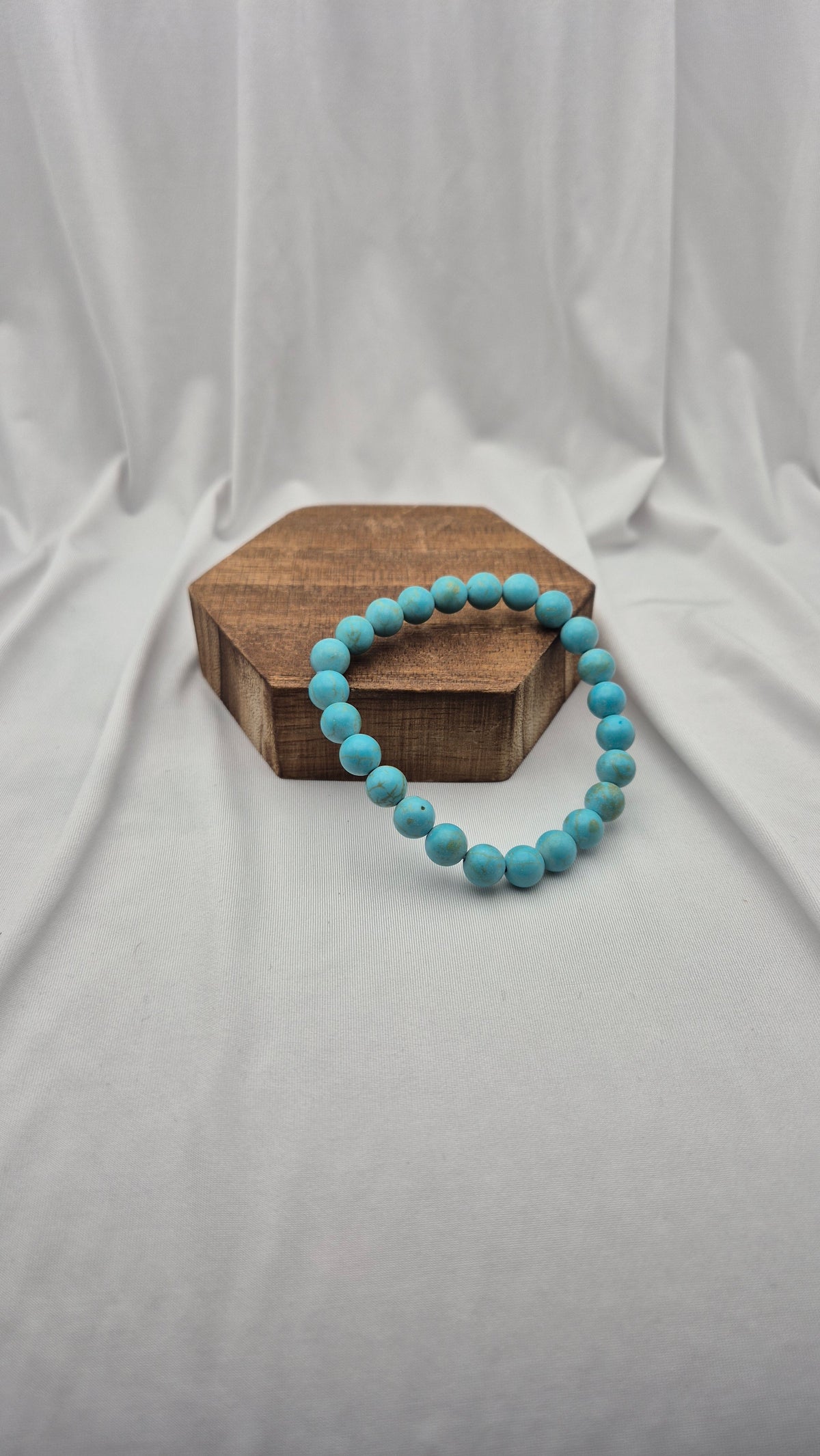 A turquoise beaded bracelet resting on a hexagonal wooden block against a draped white fabric backdrop.