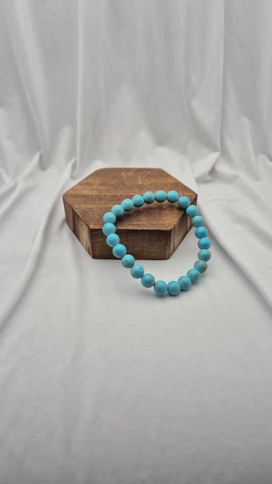 A turquoise beaded bracelet resting on a hexagonal wooden block against a draped white fabric backdrop.