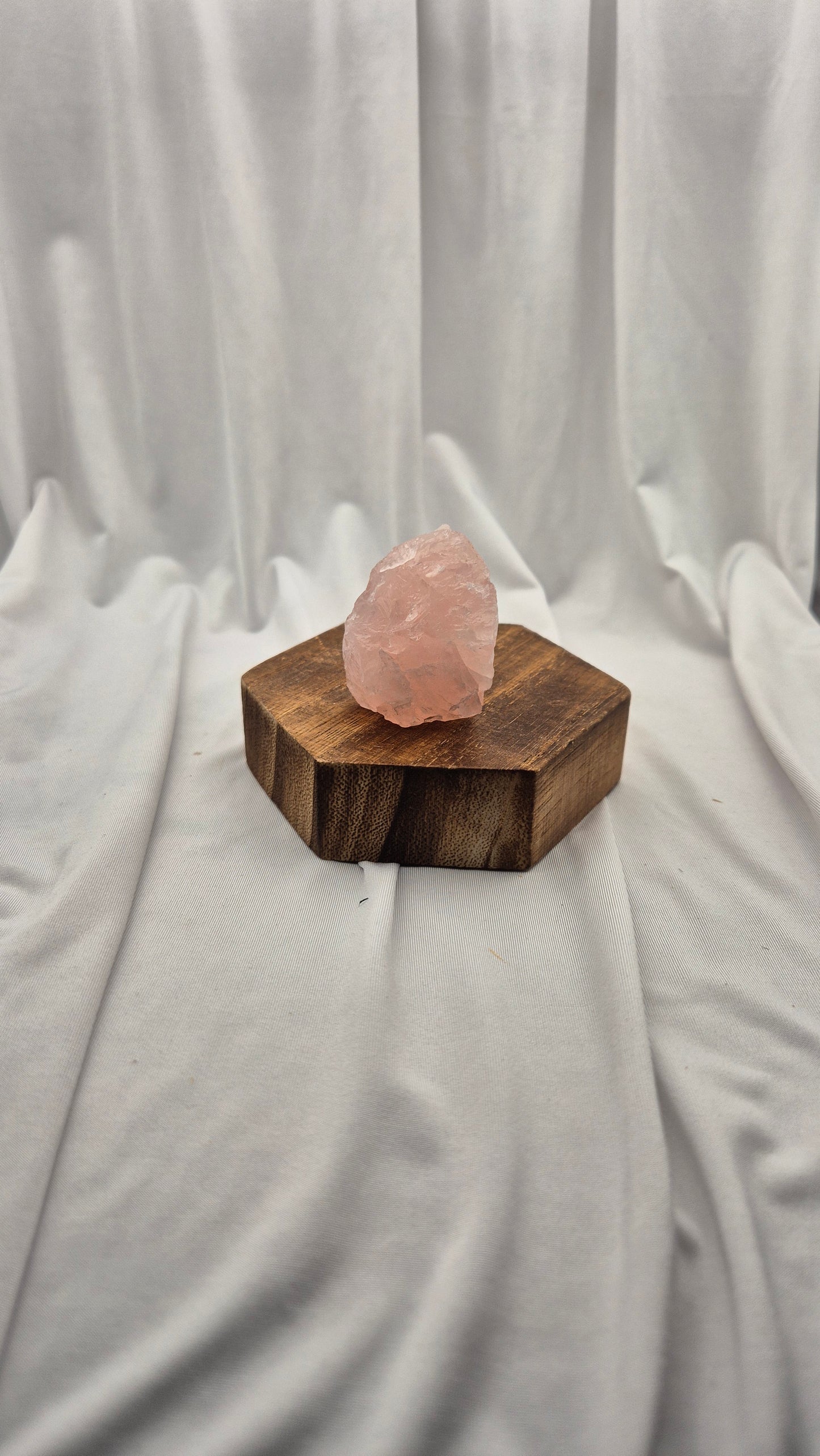 A raw pink quartz crystal resting on a hexagonal dark brown wooden block, set against a draped white fabric backdrop.