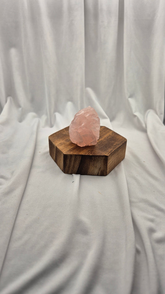 A raw pink quartz crystal resting on a hexagonal dark brown wooden block, set against a draped white fabric backdrop.