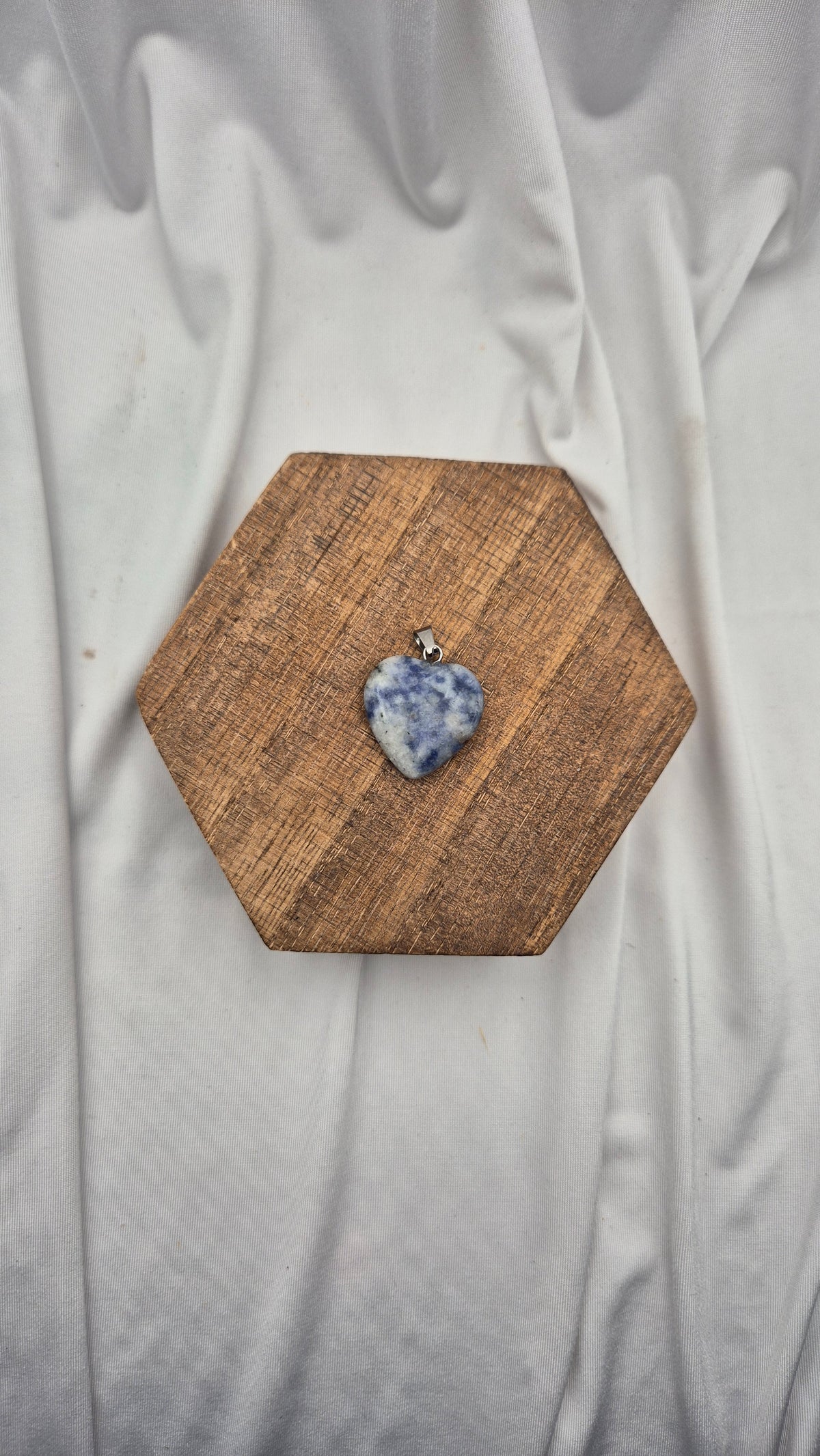 A small heart-shaped blue and white marbled stone pendant with a silver bail, displayed on a hexagonal wooden board over a draped white fabric background.