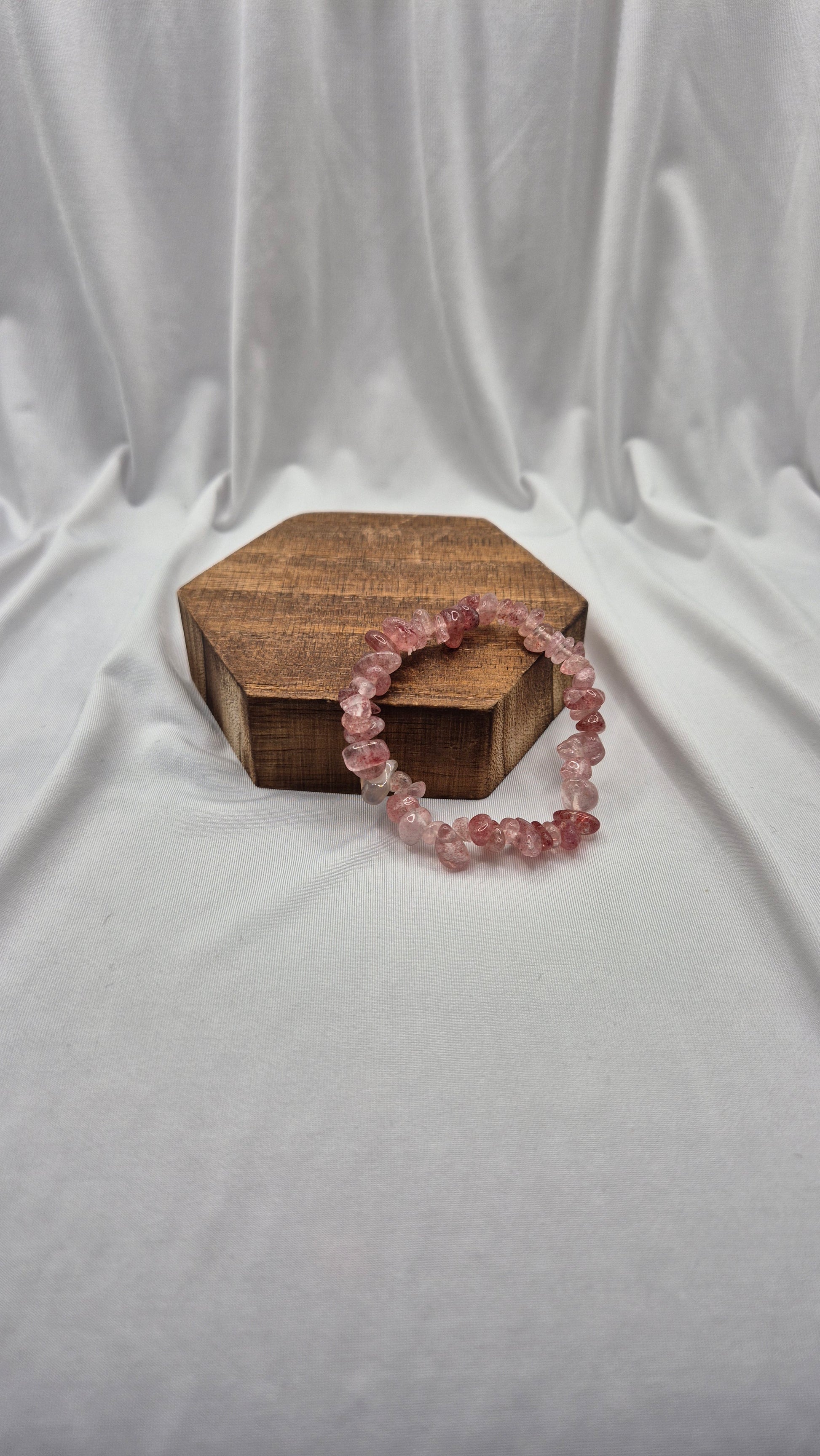 A pink beaded stretch bracelet made of faceted crystal-like beads, displayed on a small hexagonal wooden box against a draped white fabric backdrop.