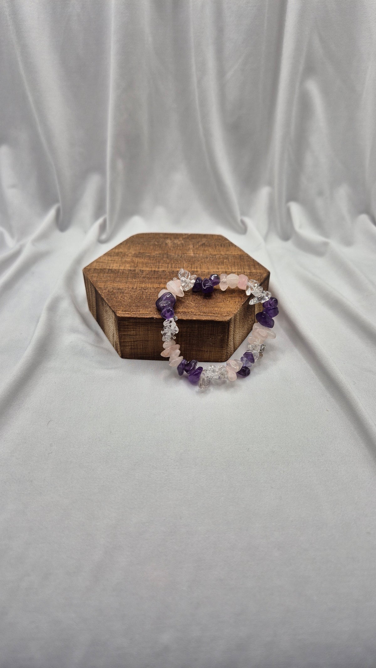 A beaded stretch bracelet with purple amethyst and light pink rose quartz beads, displayed on a small hexagonal wooden box against a draped white fabric background.