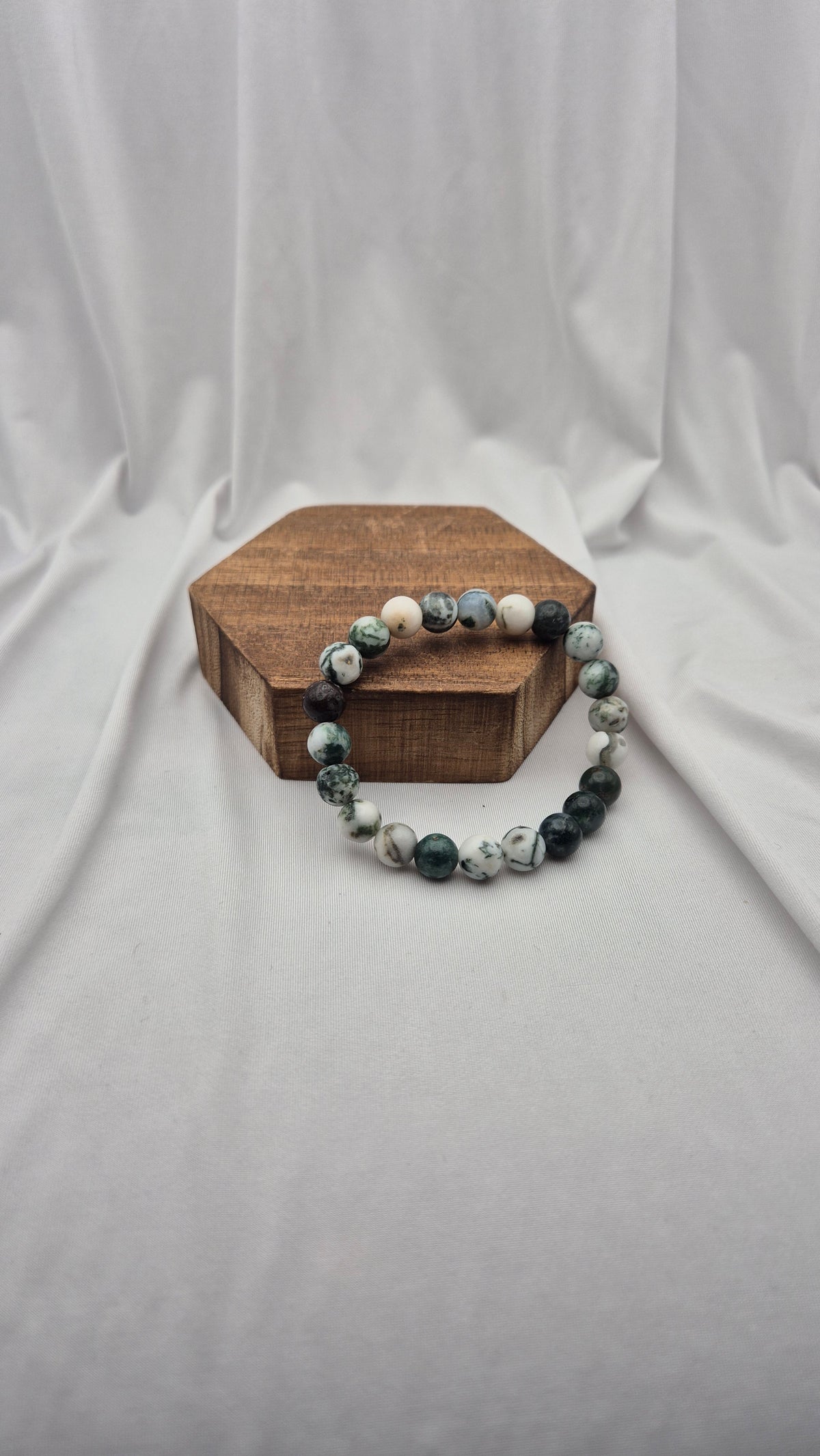 A beaded stretch bracelet made of polished round stones in shades of dark green, white, and gray, displayed on a small hexagonal wooden block against a draped white fabric backdrop.