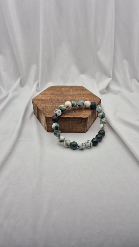 A beaded stretch bracelet made of polished round stones in shades of dark green, white, and gray, displayed on a small hexagonal wooden block against a draped white fabric backdrop.