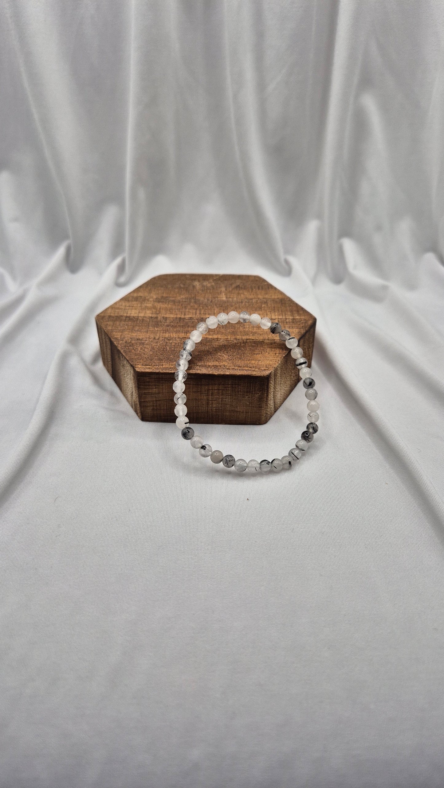 A beaded stretch bracelet made of clear and gray faceted beads, draped over a small hexagonal wooden box on a draped white fabric background.