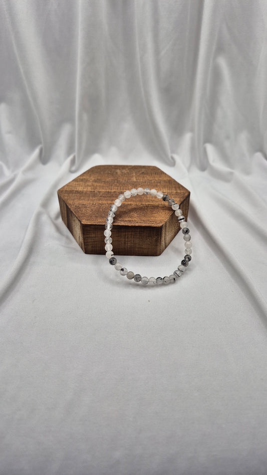 A beaded stretch bracelet made of clear and gray faceted beads, draped over a small hexagonal wooden box on a draped white fabric background.