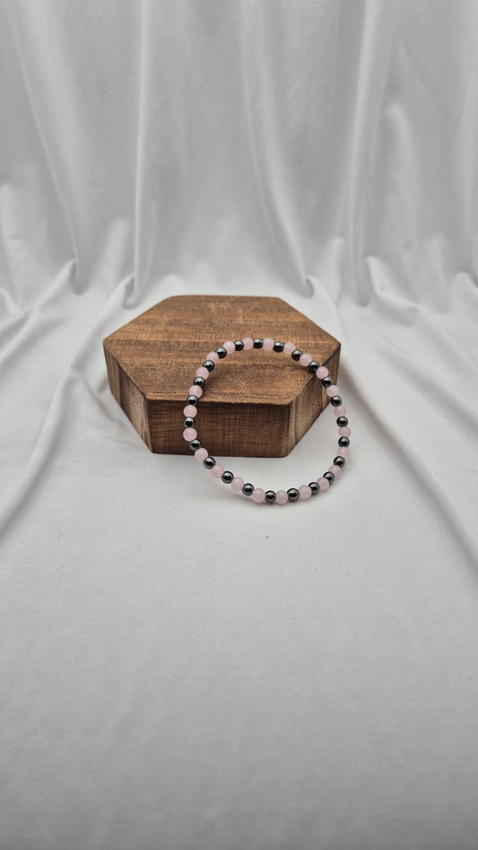 A beaded stretch bracelet with alternating black and light pink beads, displayed on a small hexagonal wooden box against a draped white fabric backdrop.