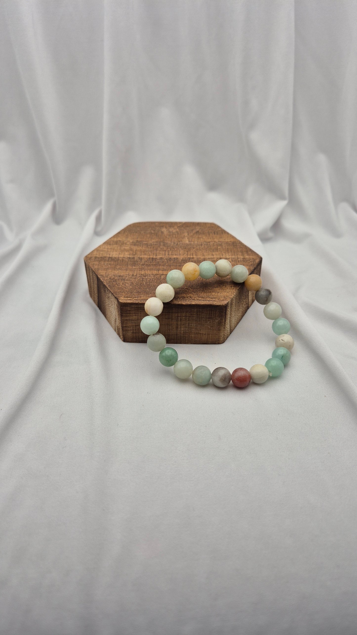 A beaded stretch bracelet made of round multicolor gemstone-like beads, displayed on a small hexagonal wooden block against a draped white fabric backdrop.
