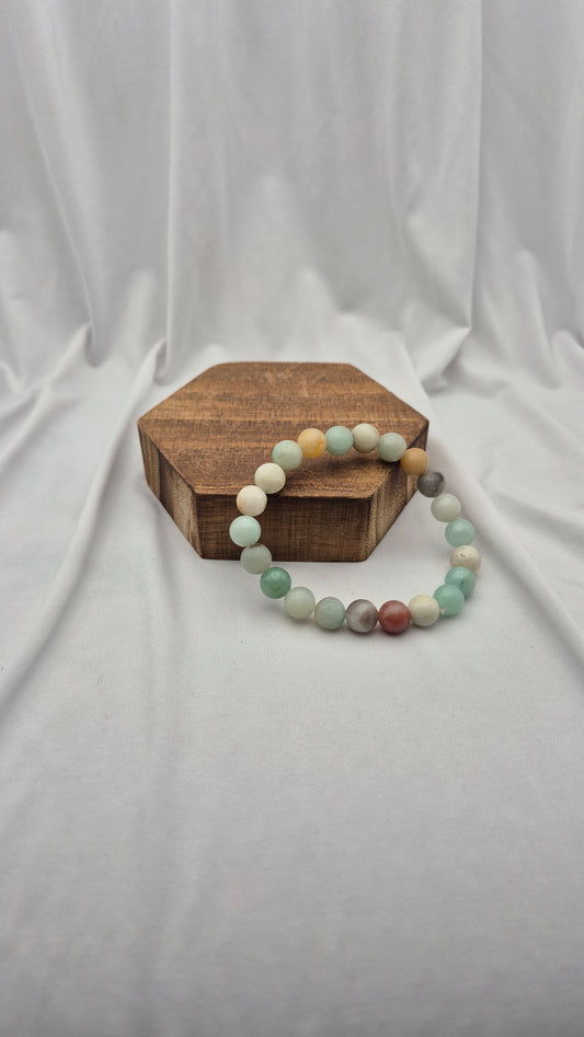 A beaded stretch bracelet made of round multicolor gemstone-like beads, displayed on a small hexagonal wooden block against a draped white fabric backdrop.