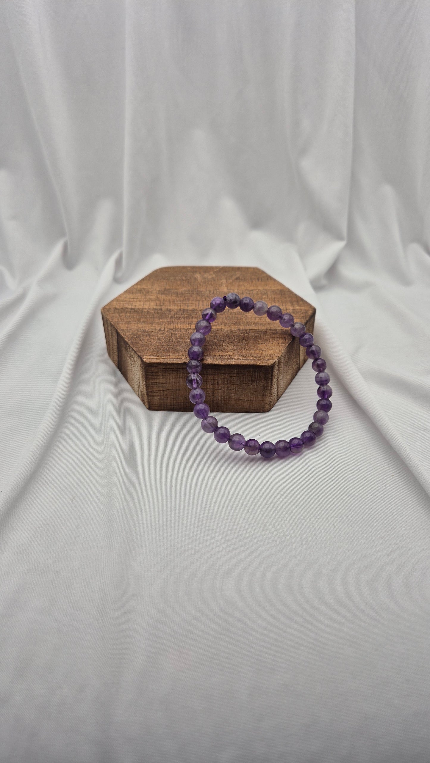 A purple beaded bracelet resting on a small hexagonal wooden block against a draped white fabric backdrop.