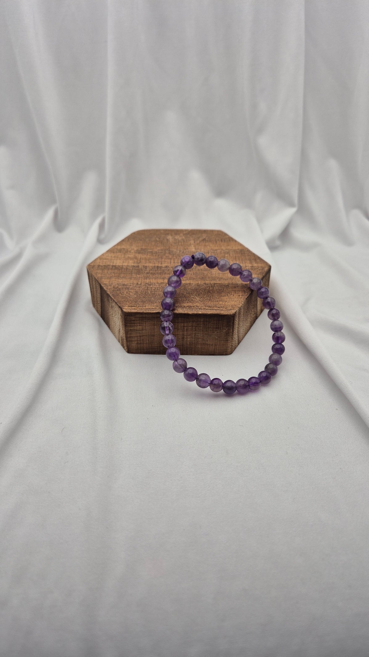 A purple beaded bracelet resting on a small hexagonal wooden block against a draped white fabric backdrop.