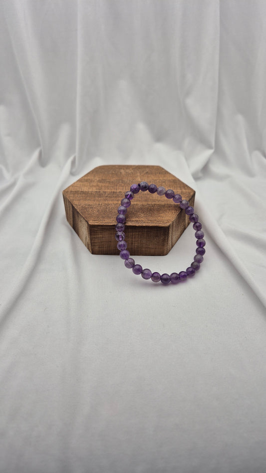 A purple beaded bracelet resting on a small hexagonal wooden block against a draped white fabric backdrop.