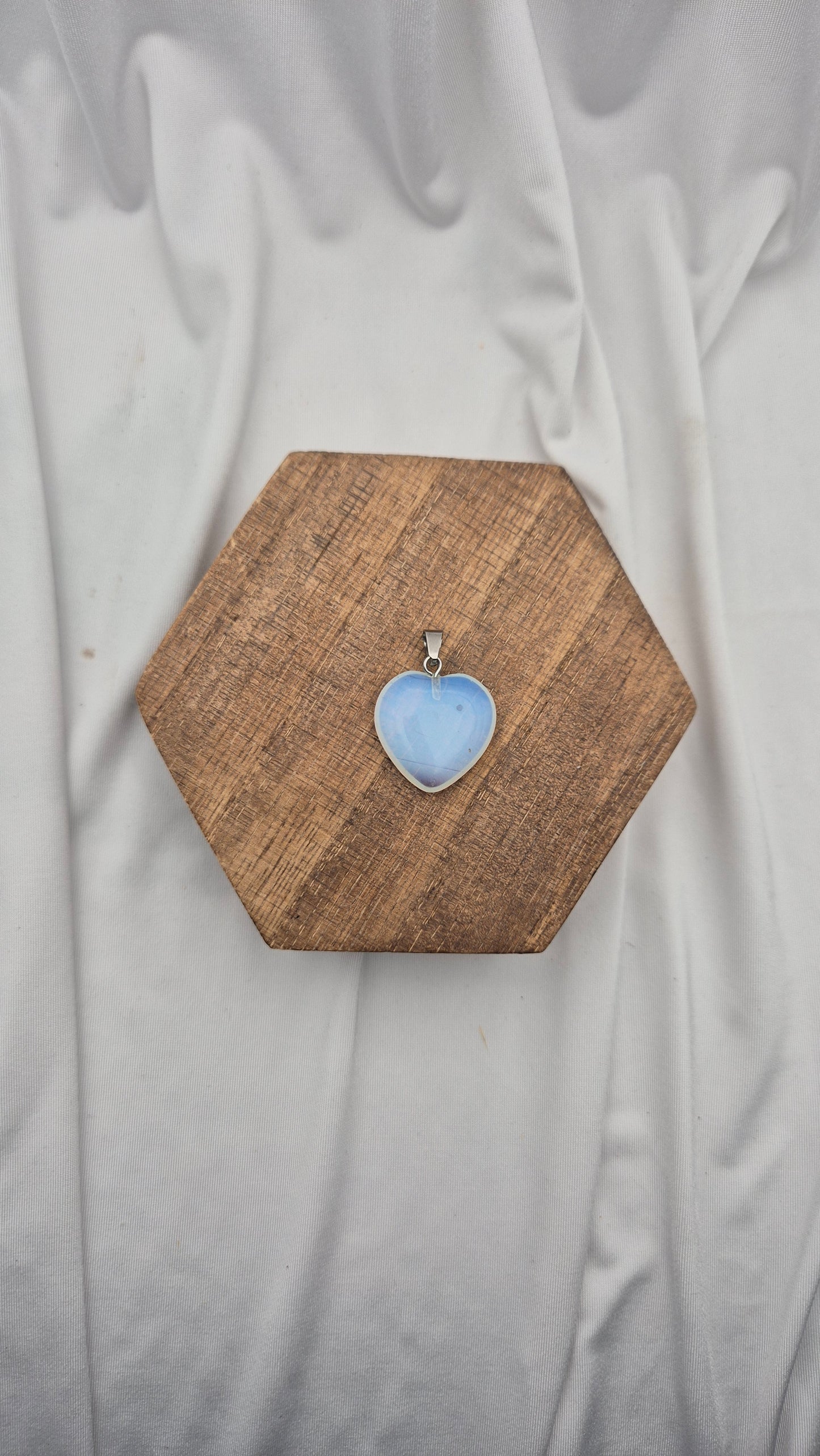 A heart-shaped light blue stone pendant in a silver bezel setting, displayed on a hexagonal wooden block over a draped white fabric background.