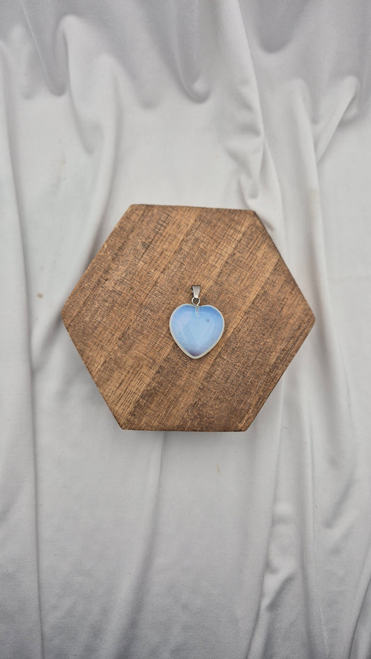 A heart-shaped light blue stone pendant in a silver bezel setting, displayed on a hexagonal wooden block over a draped white fabric background.