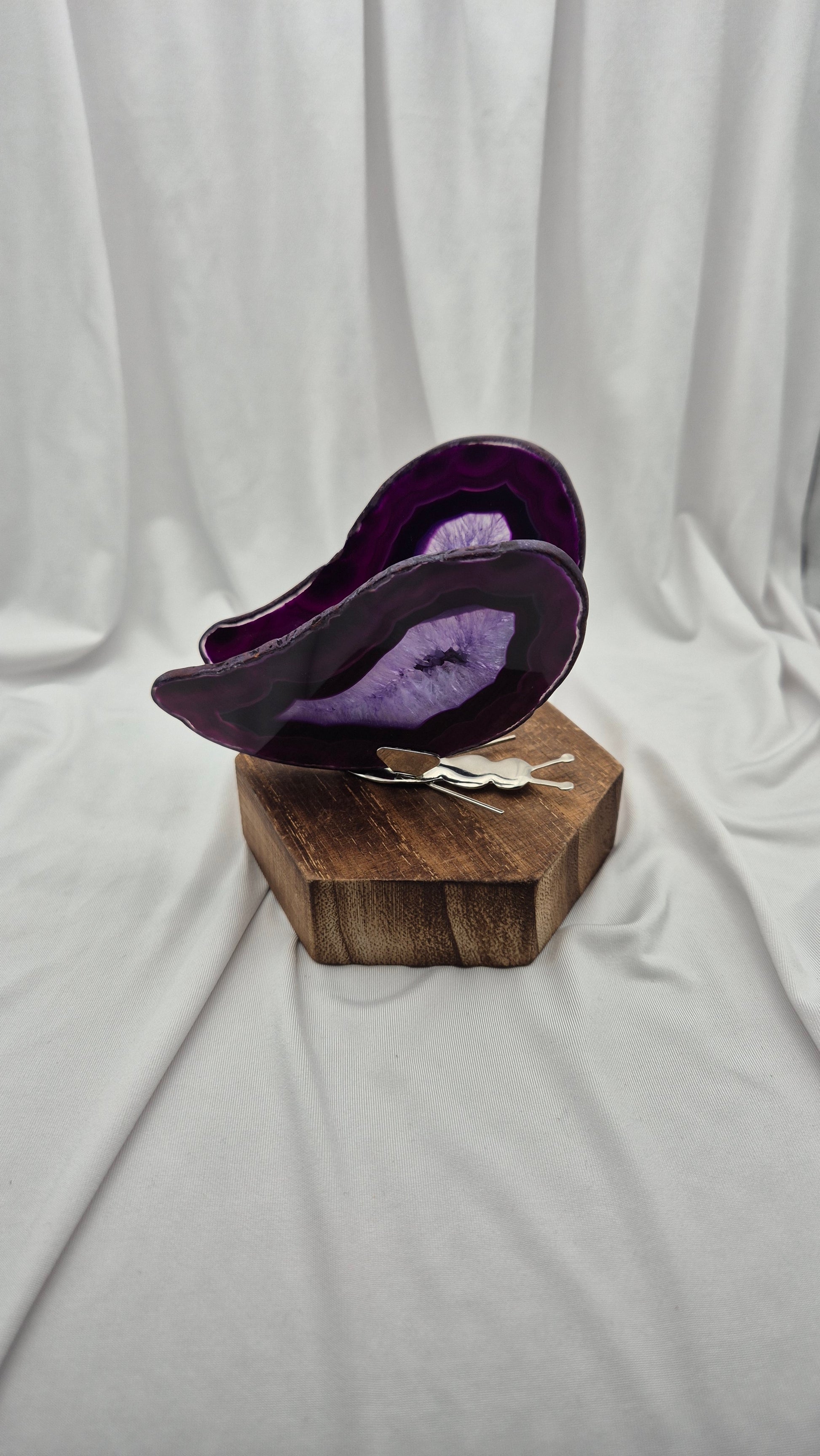 A purple agate slice decorative bowl displayed on a hexagonal wooden block stand, with a small white tag attached, set against a draped white fabric backdrop.